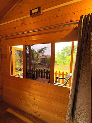 A wooden cabin with a window overlooking a garden and a red-roofed building. - Hill View Paradise (Gokarna)