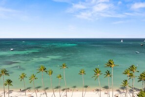 Una playa con aguas turquesas cristalinas, palmeras y un barco a lo lejos.