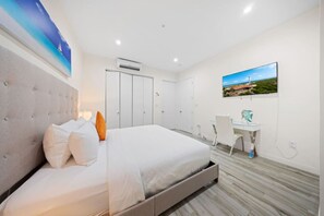 Quarto de hotel com cama, escrivaninha e uma cadeira. Há um quadro na parede e um ar condicionado sobre a cama.