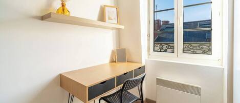 A small, bright room with a wooden desk, a chair, a radiator, and a window with a view of buildings.