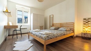 A bedroom with a wooden bed, a desk with a chair, a white rug, and a view of the outside through a window. - Charming Bright Duplex (Angers)