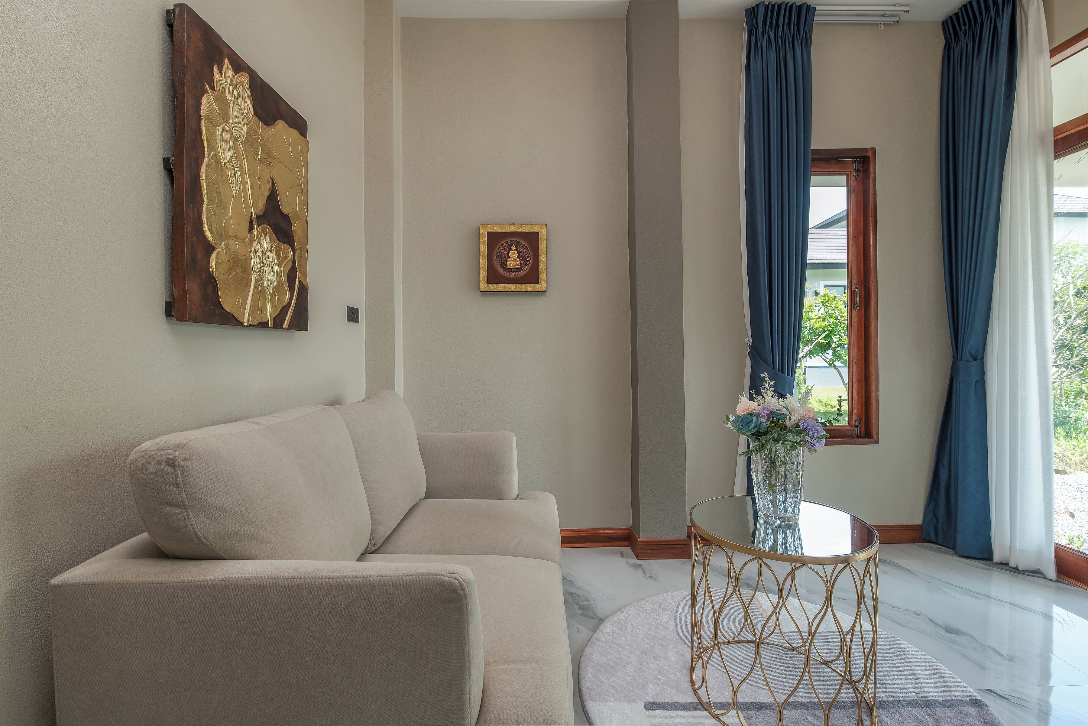 A living room with a beige sofa, a glass table with a gold frame, and a vase with flowers.