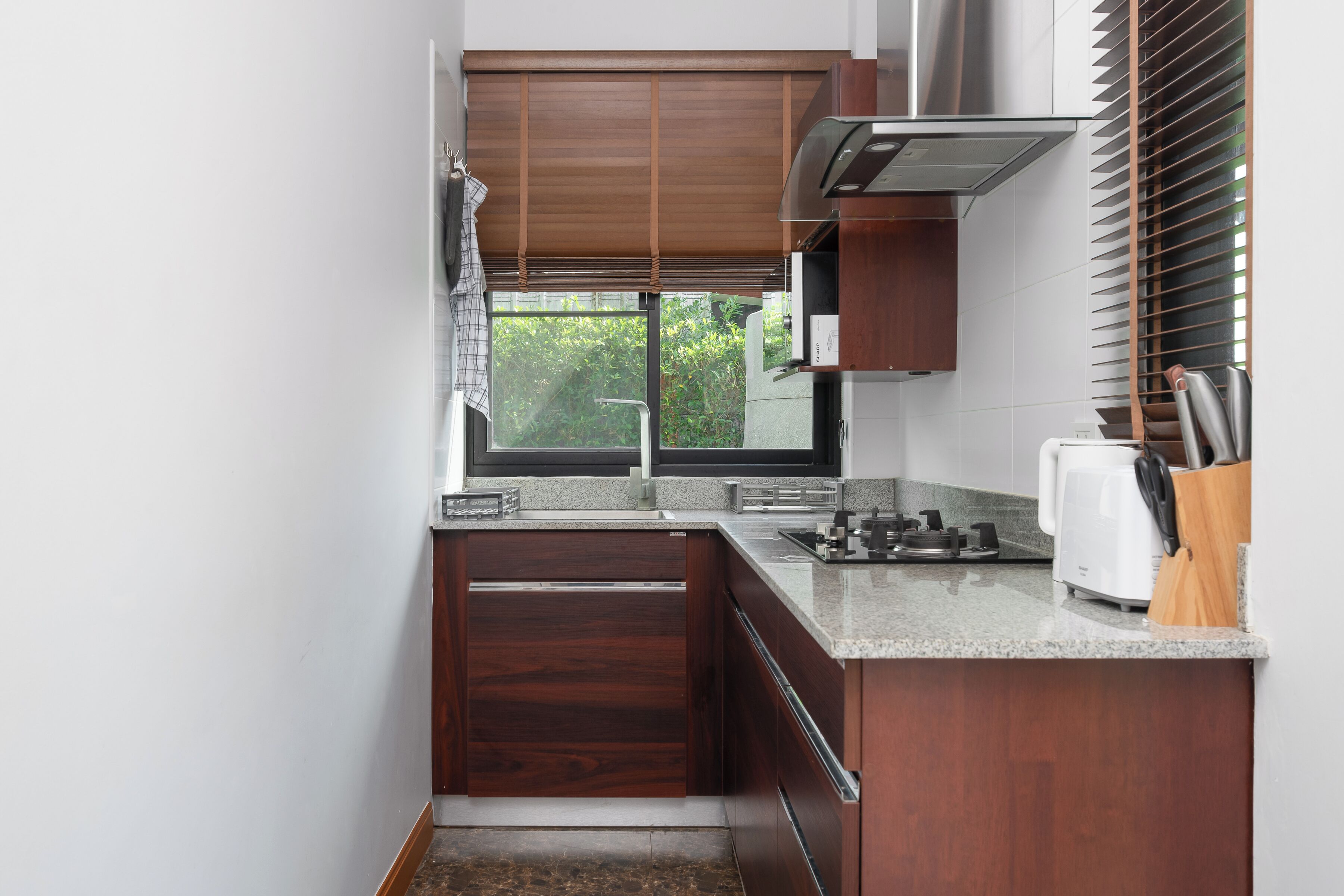 A compact kitchen with dark wood cabinets, a granite countertop, a built-in stove, and a window with blinds.