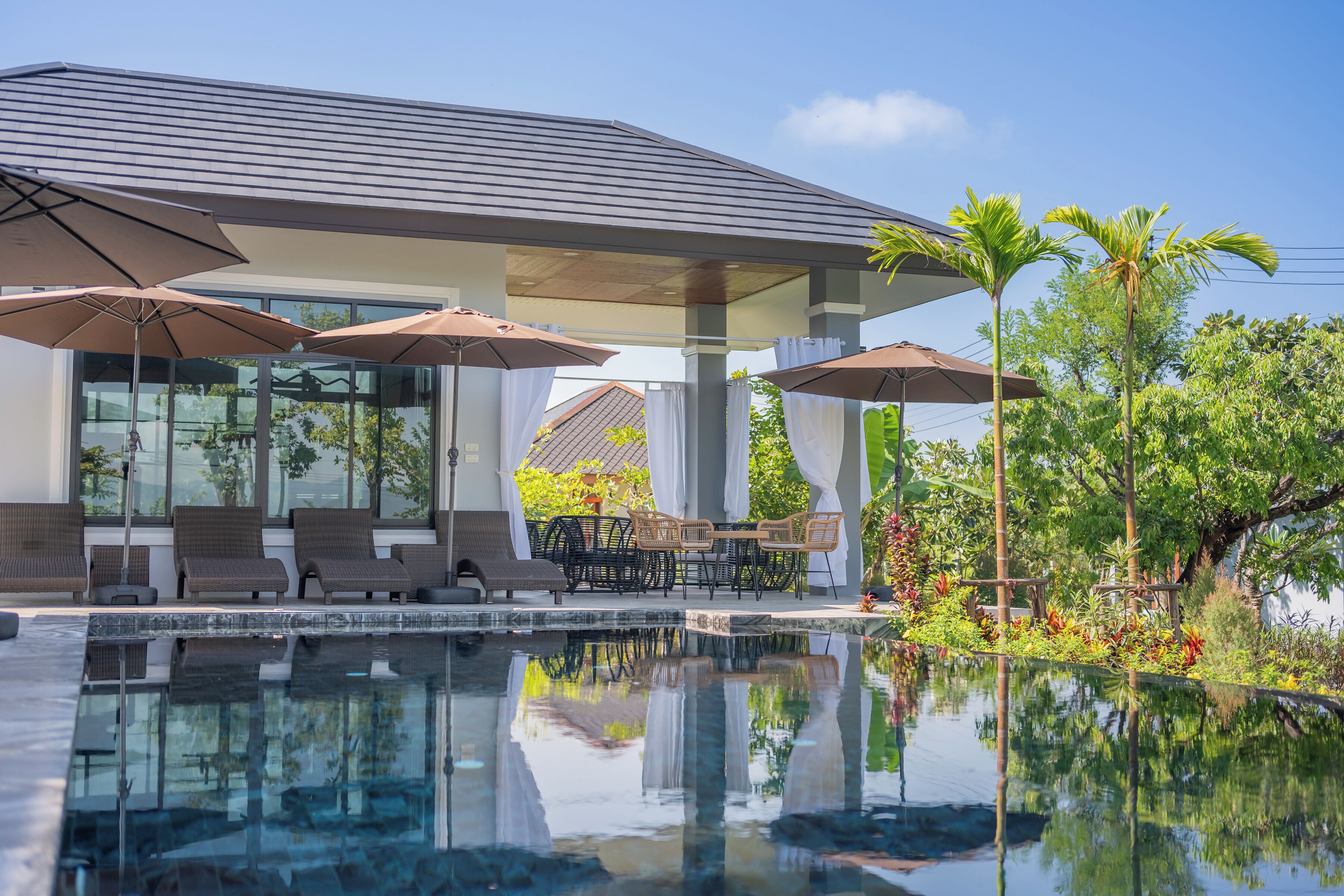 Un área de piscina moderna con sillones de descanso y sombrillas, rodeada de exuberante vegetación y un cielo despejado.