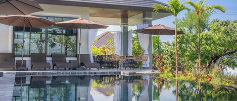 Un área de piscina moderna con sillones de descanso y sombrillas, rodeada de exuberante vegetación y un cielo despejado.