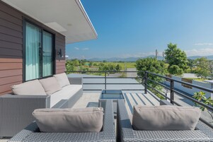 Un balcón con mobiliario de mimbre, una mesa de vidrio y vistas a un paisaje verde y montañas.