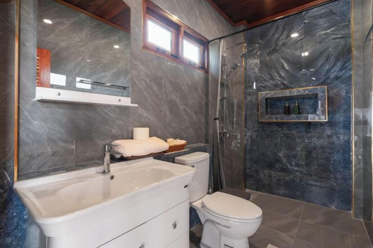 Un baño con pared de mármol, lavamanos blanco, inodoro y ducha con mampara de vidrio.