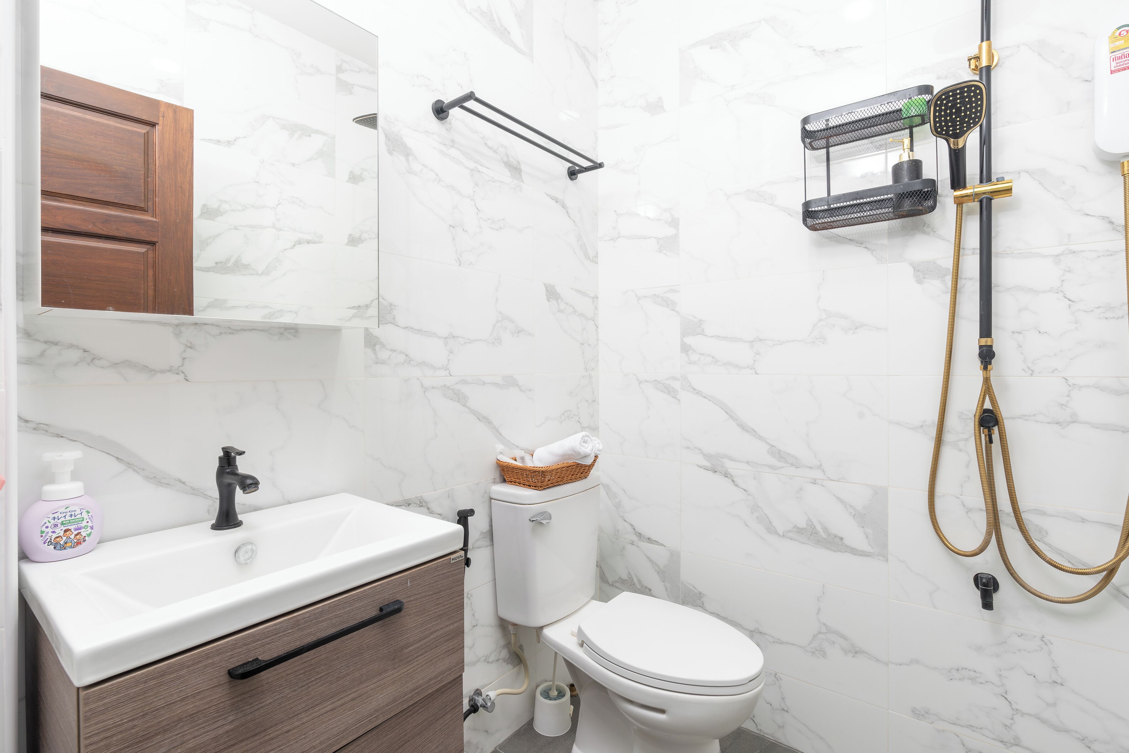 Un baño con pared de mármol, un lavamanos blanco, un armario de madera, un inodoro y una ducha con manguera.