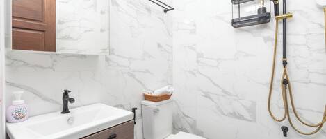Un baño con pared de mármol, un lavamanos blanco, un armario de madera, un inodoro y una ducha con manguera.