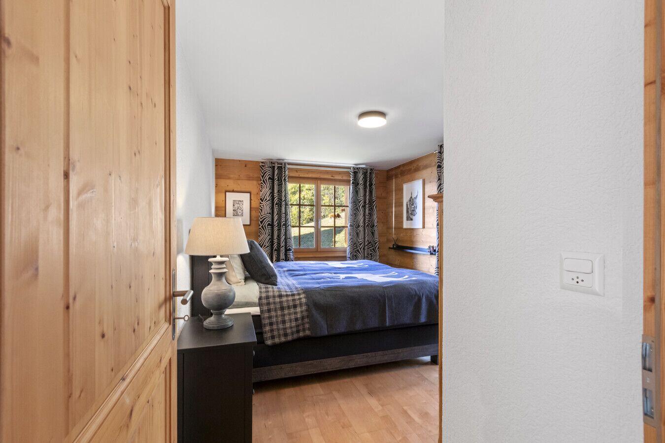 A bedroom with a bed, a bedside table with a lamp, a window with curtains, and wooden walls.