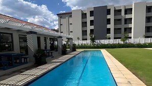 A swimming pool with a tiled deck, a covered seating area, and a building with multiple balconies in the background. - The Henlee Apartment Tocara (Chartwell)