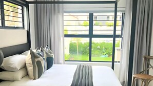 A modern bedroom with a bed, curtains, and a view of greenery outside. - The Henlee Apartment Tocara (Chartwell)