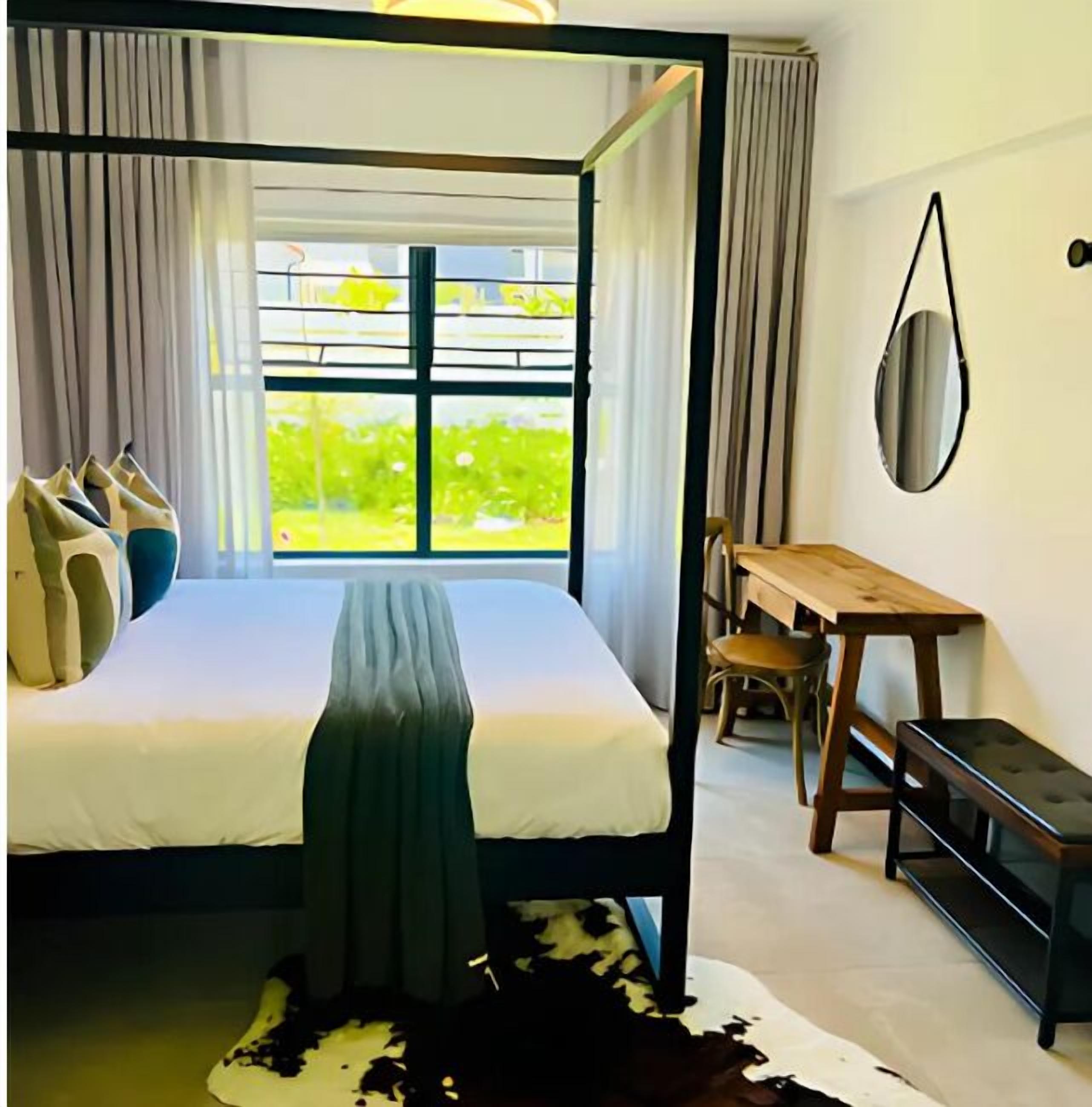 A four-poster bed with a view of greenery outside, a wooden desk, and a mirror on the wall.