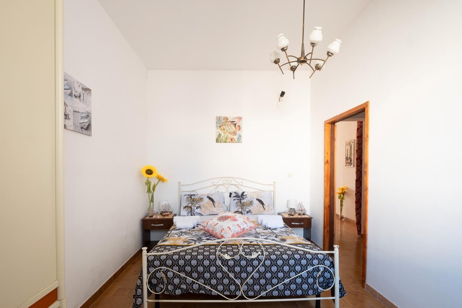 A bedroom with a bed, a chandelier, a nightstand, a vase with sunflowers, and a door.