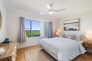 Une chambre avec un grand lit, un ventilateur de plafond, une fenêtre avec des rideaux, une table de chevet avec une lampe et un tableau au mur.