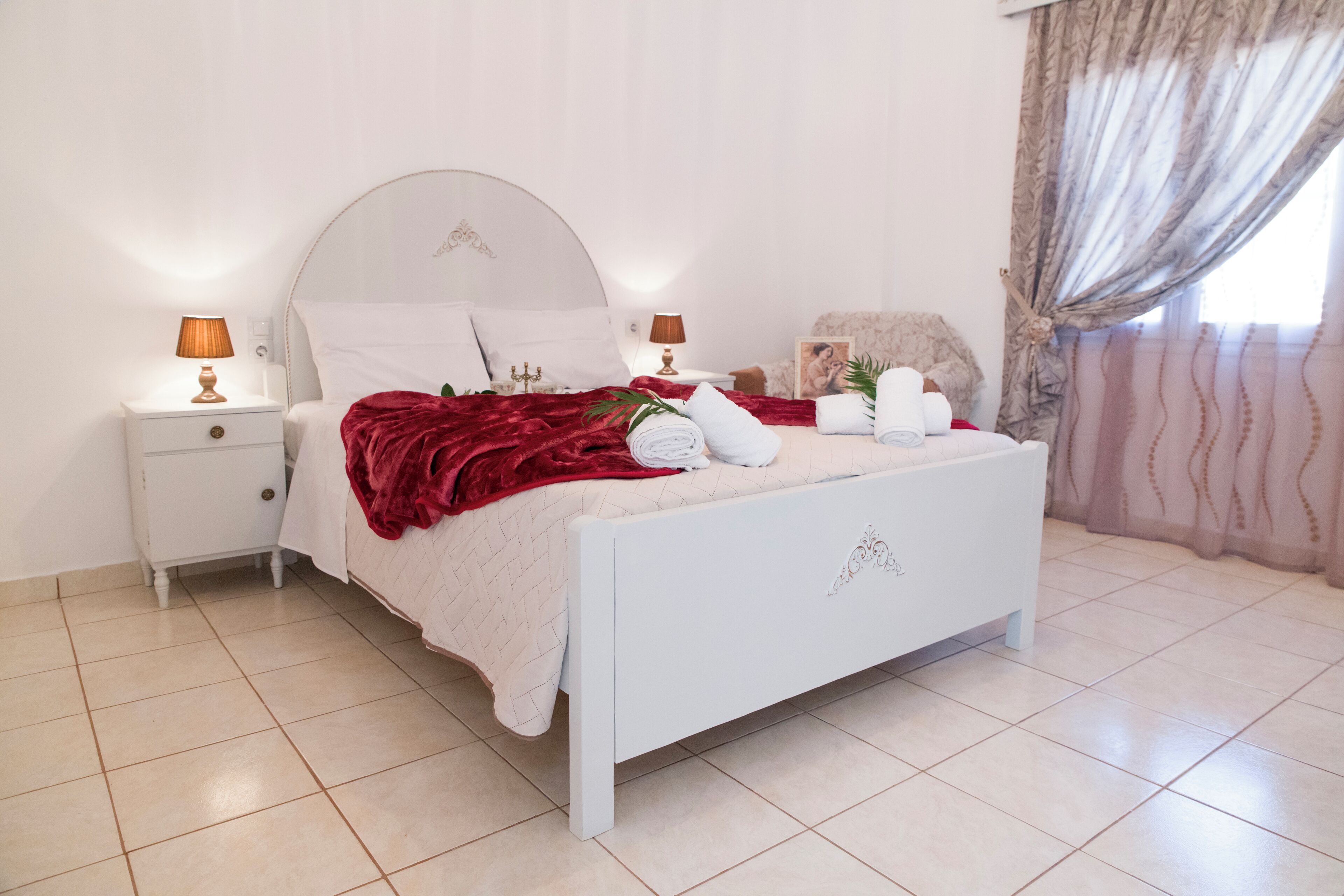 Un lit bien fait, avec une tête de lit blanche, un plaid rouge et des serviettes soigneusement pliées. Il y a deux tables de chevet avec des lampes, et une fenêtre ornée de rideaux légers.