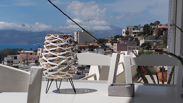 Een balkon met een tafel en stoelen, met uitzicht op een kustplaatsje met gebouwen en een berg op de achtergrond.