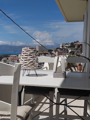 A balcony with a table and chairs, overlooking a coastal town with buildings and a mountain in the background. - CHRYSO APARTMENT (Kamilari)