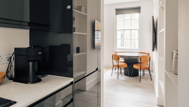 A modern kitchen with a coffee maker, a dining area with a round table and orange chairs, and a large window with blinds. - Stylish newly refurbished 2-bed retreat in the heart of London (London)