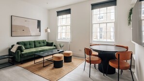 A modern living room with a green sofa, a round dining table with chairs, and large windows with blinds. - Stylish newly refurbished 2-bed retreat in the heart of London (London)