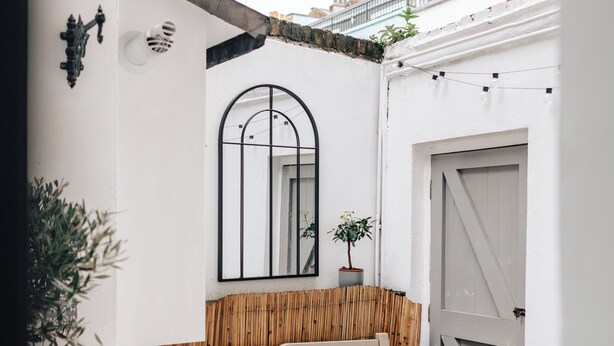 Un piccolo spazio esterno recintato, con una panchina in legno, un muro bianco con uno specchio e una porta.