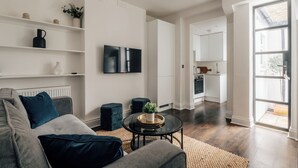 Un soggiorno moderno con un divano grigio, un poggiapiedi nero e un tavolino in vetro. C'è una TV a schermo piatto montata sulla parete, una pianta in vaso e uno scaffale con oggetti decorativi. Si intravede una cucina sullo sfondo.