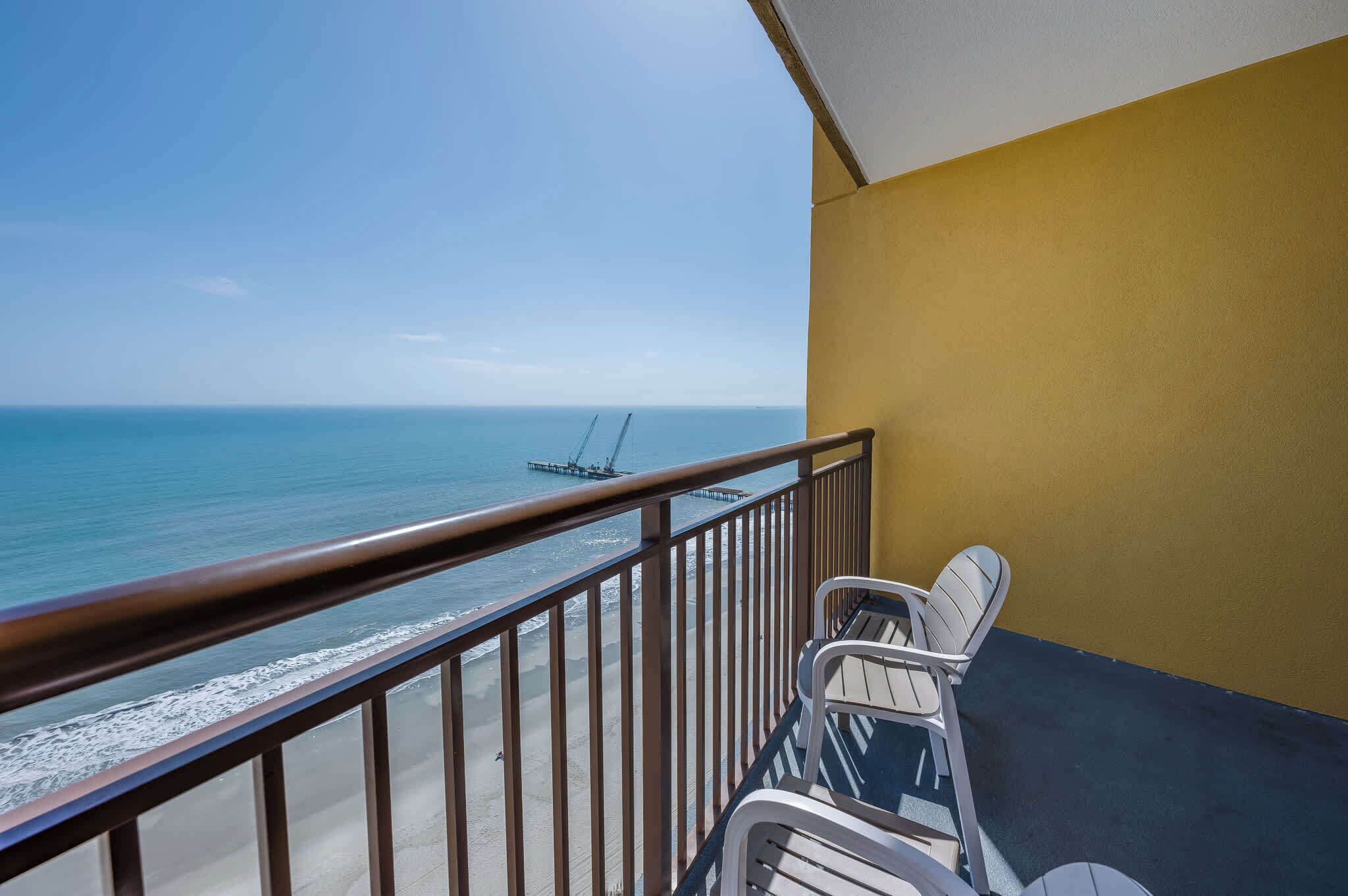 Ein Balkon mit Blick auf das Meer und einen Steg. Zwei weiße Stühle und ein Tisch befinden sich auf dem Balkon.