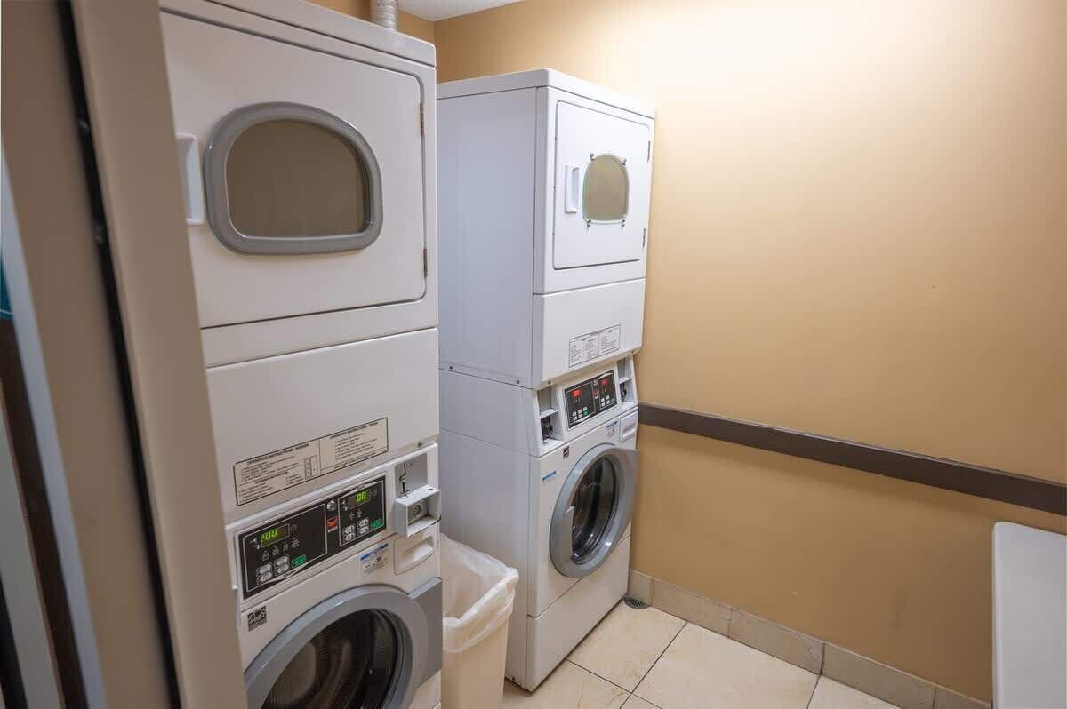 Un cuarto de lavandería con lavadoras y secadoras apiladas, un panel de control y un bote de basura.