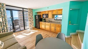 A modern kitchen with wooden cabinets, a refrigerator, and a microwave. There is a dining area with a table and chairs, and a balcony with a view of the ocean. - Direct Oceanfront 1 BR, Warm Swimming Pools,Views! (North Myrtle Beach)