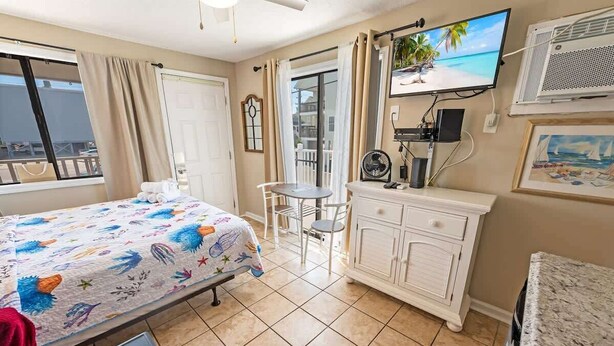 A hotel room with a bed, a small table, a TV, a fan, and a view of the beach through the window.