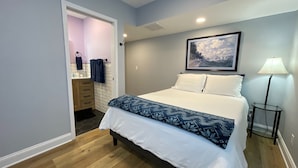 A bedroom with a bed, a bedside lamp, a painting on the wall, and a bathroom visible through an open door.