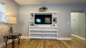 A living room with a flat-screen TV mounted on a white entertainment unit, a glass coffee table with a metal frame, and a lamp on a side table.