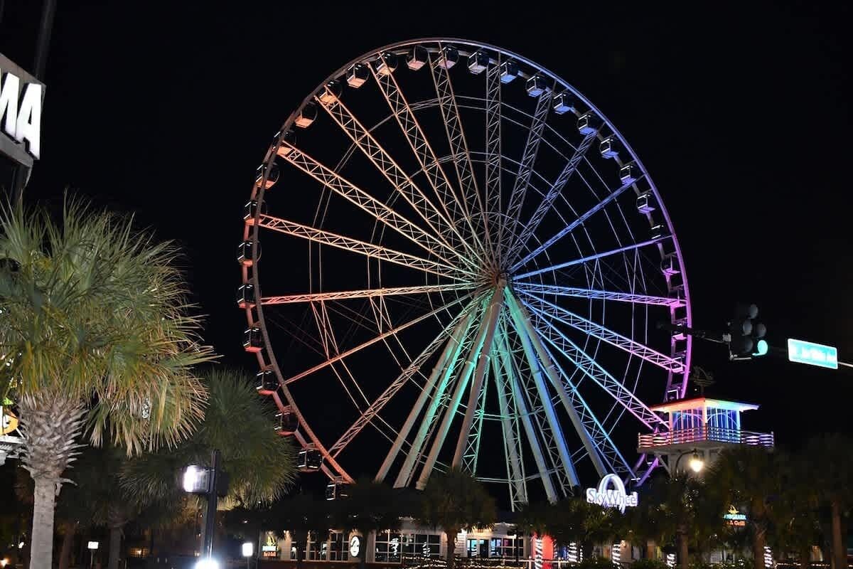 A large Ferris wheel with multiple cabins, illuminated with colorful lights, and a building with a sign that reads "SkyWheel" in the foreground.