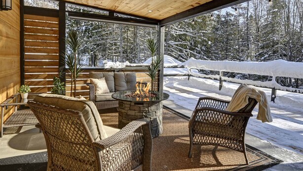 A cozy outdoor seating area with wicker chairs and a stone fire pit, overlooking a snowy landscape with trees and a bench covered in snow. - Refuge Tremblant Spa Ski 11min (Lac-Supérieur)