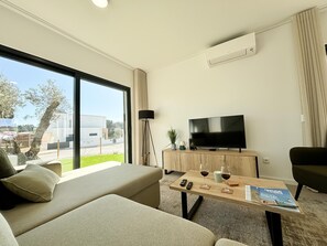Habitación de hotel moderna con sofá, una mesita de centro con una revista, televisión y un ventanal con vistas al exterior.