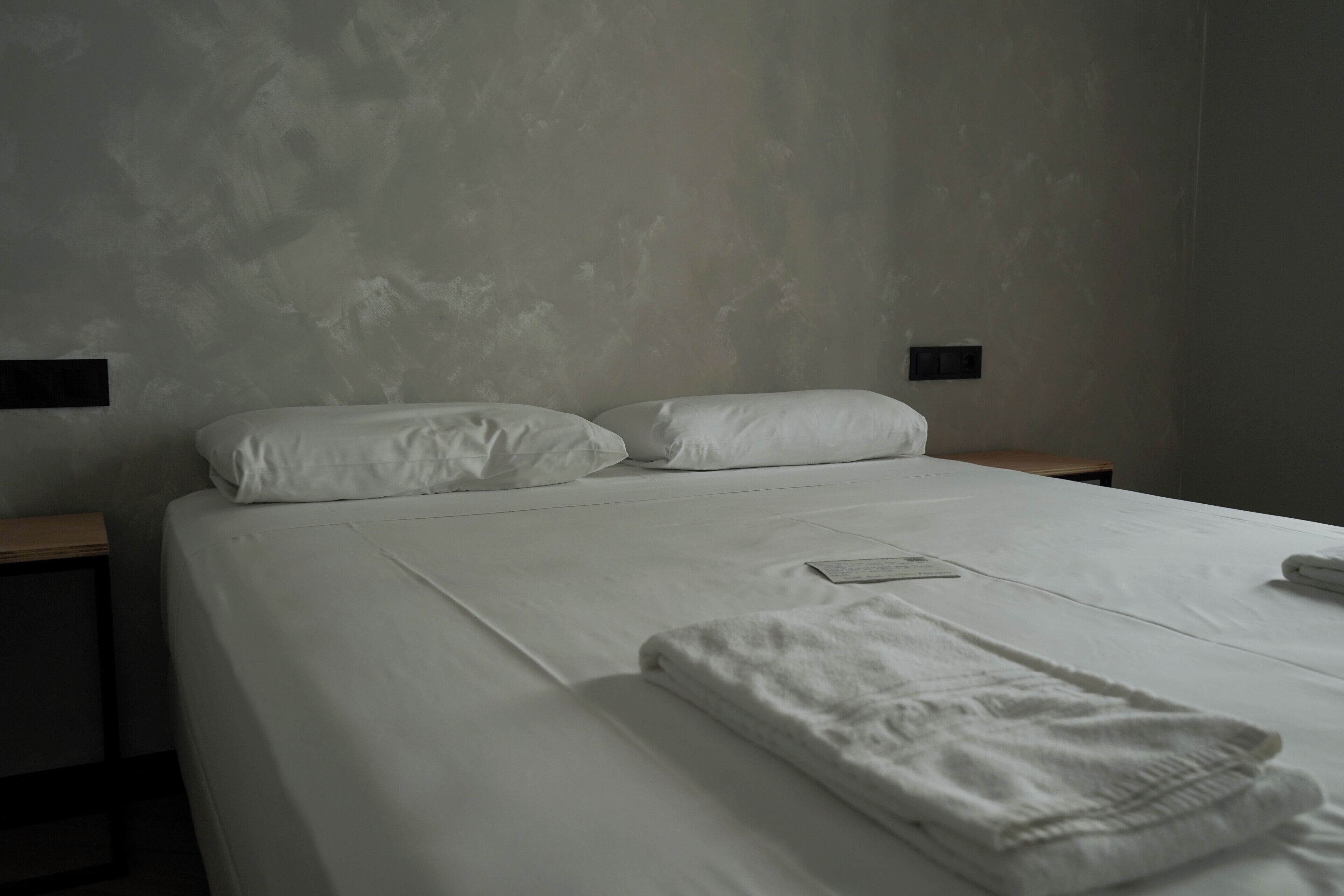 A neatly made bed with white linens, a folded towel, and a small bedside table with a lamp.