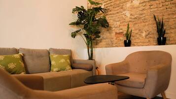 Un salón con un sofá, un sillón y una mesita. Hay tres plantas en macetas junto a una pared de ladrillo.