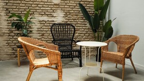 A room with two wicker chairs and a small round table, a brick wall, and large green plants. - Sweet Sevilla Hostel  (Seville)
