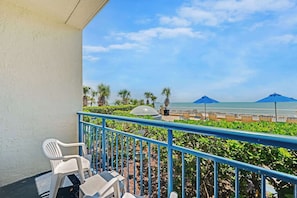 Un balcon avec des chaises et une table en plastique blanc, donnant sur une plage parsemée de parasols et de palmiers.