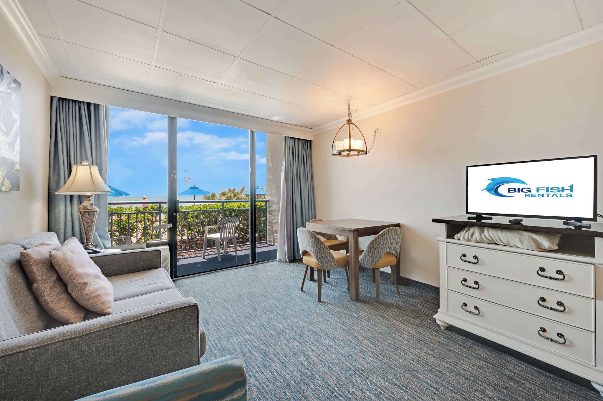 Ein Hotelzimmer mit einer Couch, einem Esstisch mit Stühlen, einem Fernseher, einem Schminktisch und einem Balkon mit Meerblick.
