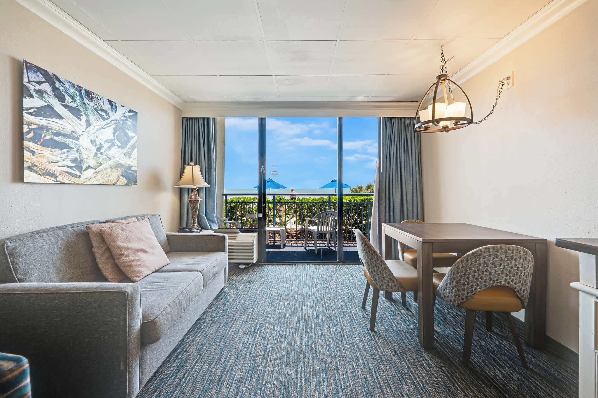 Habitación de hotel con sofá, escritorio, silla, lámpara y un ventanal con vistas a zonas verdes y montañas.