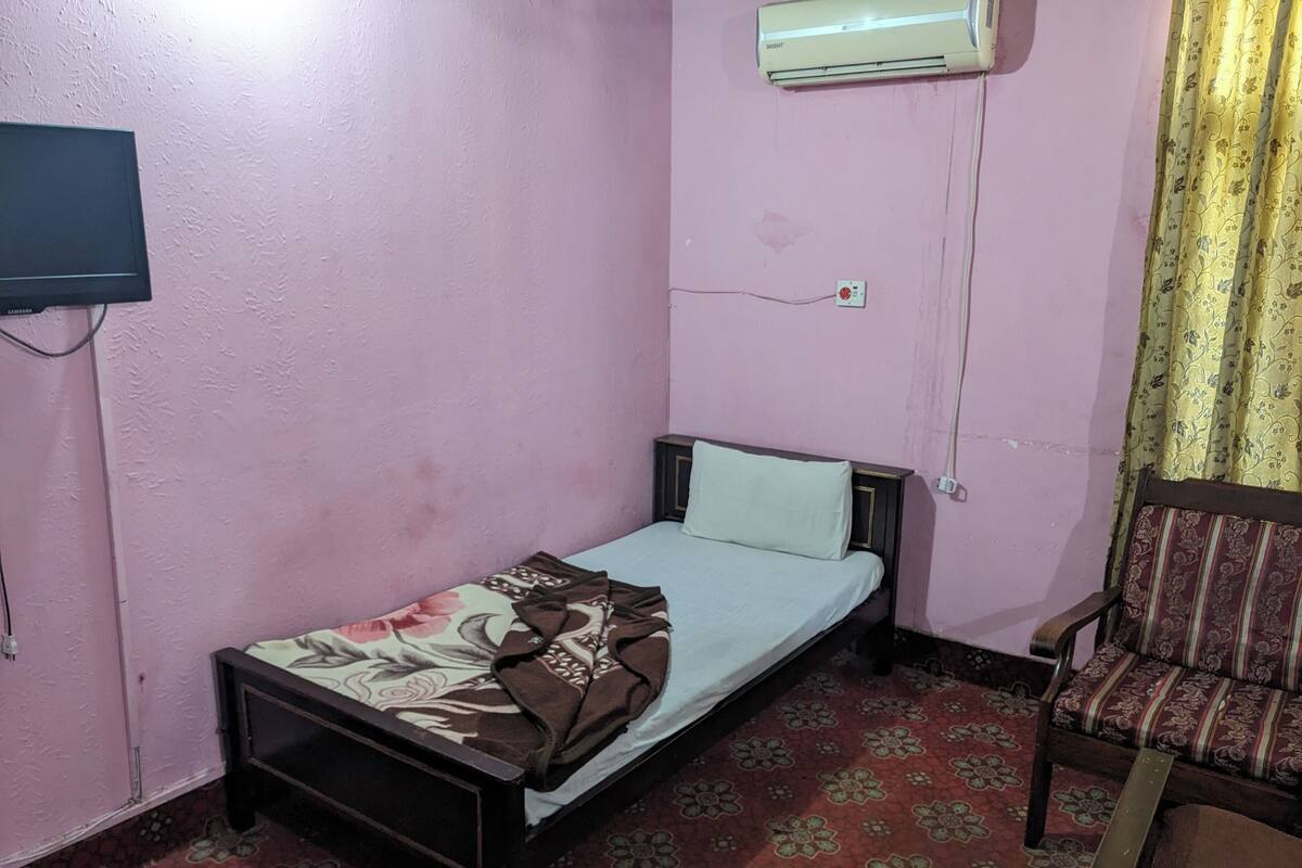 Une petite chambre avec un lit simple, un mur rose, une moquette à motifs, un téléviseur sur le côté gauche et un climatiseur au-dessus du lit.