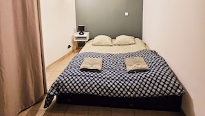 Un dormitorio con cama, almohadas, mesita de noche, lámpara y cortina.