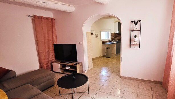 Una sala de estar con un sofá, una mesa de centro y un televisor. Se ve una cocina a través de un arco.
