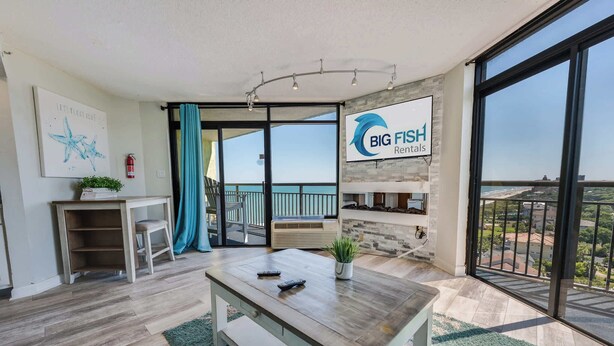 A modern living room with a large window, a fireplace, a dining area, and a view of the ocean.