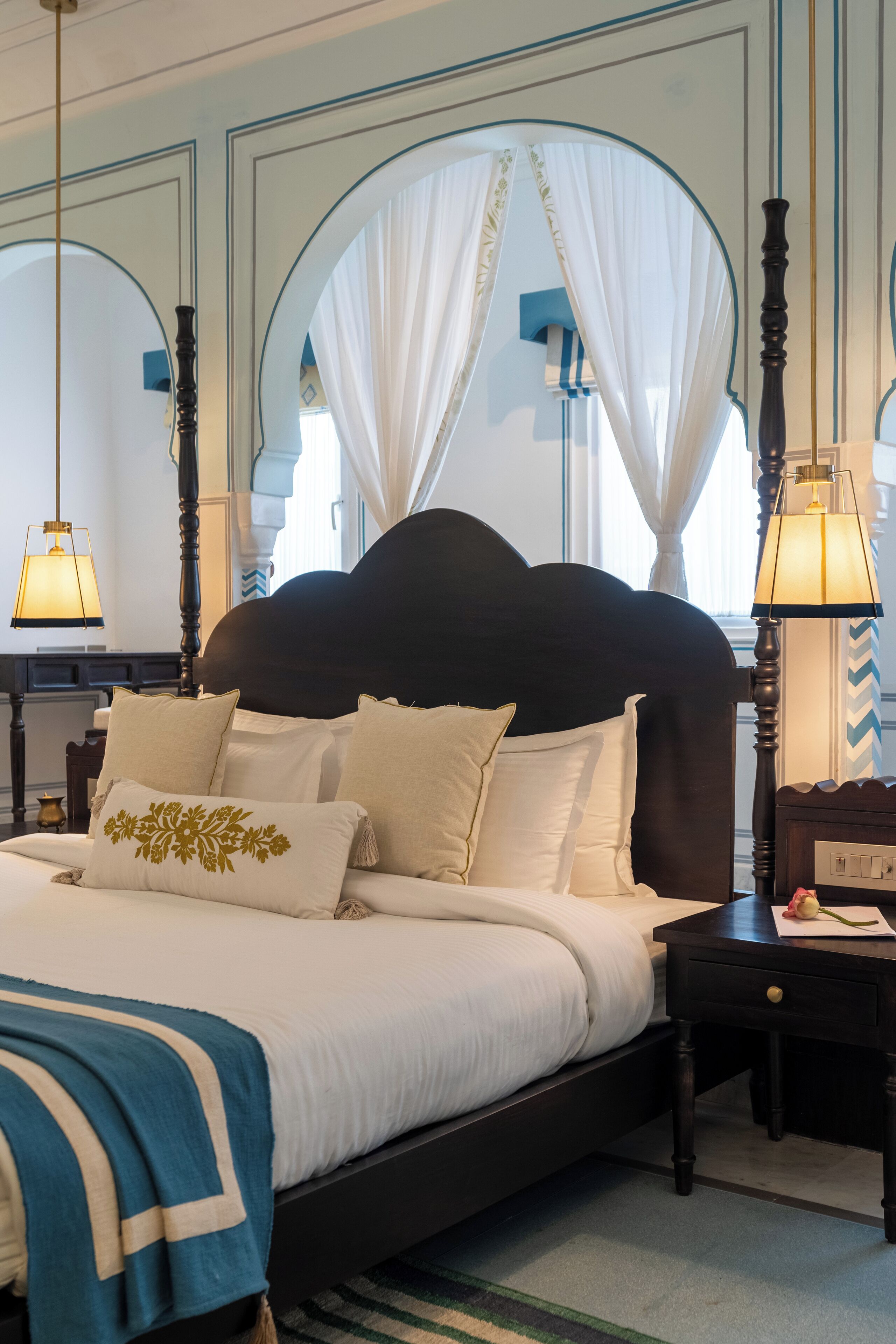 A neatly made bed with a dark headboard, white bedding, and decorative pillows. There is a bedside table with a lamp and a small decorative item. The room has a classic design with arched windows and a striped rug.