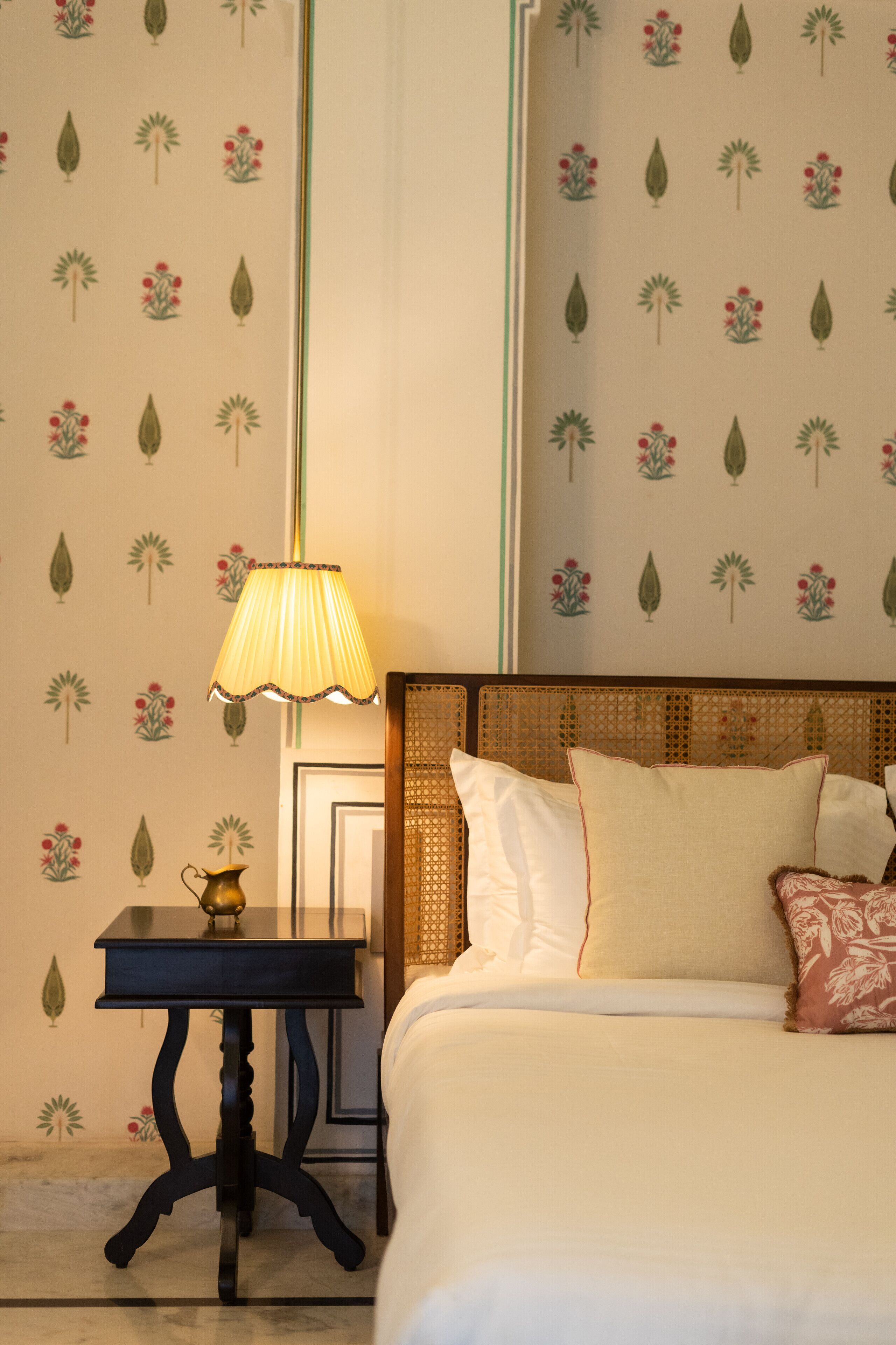 A neatly made bed with white linens and a headboard, a bedside table with a lamp, and a wall with a floral pattern.