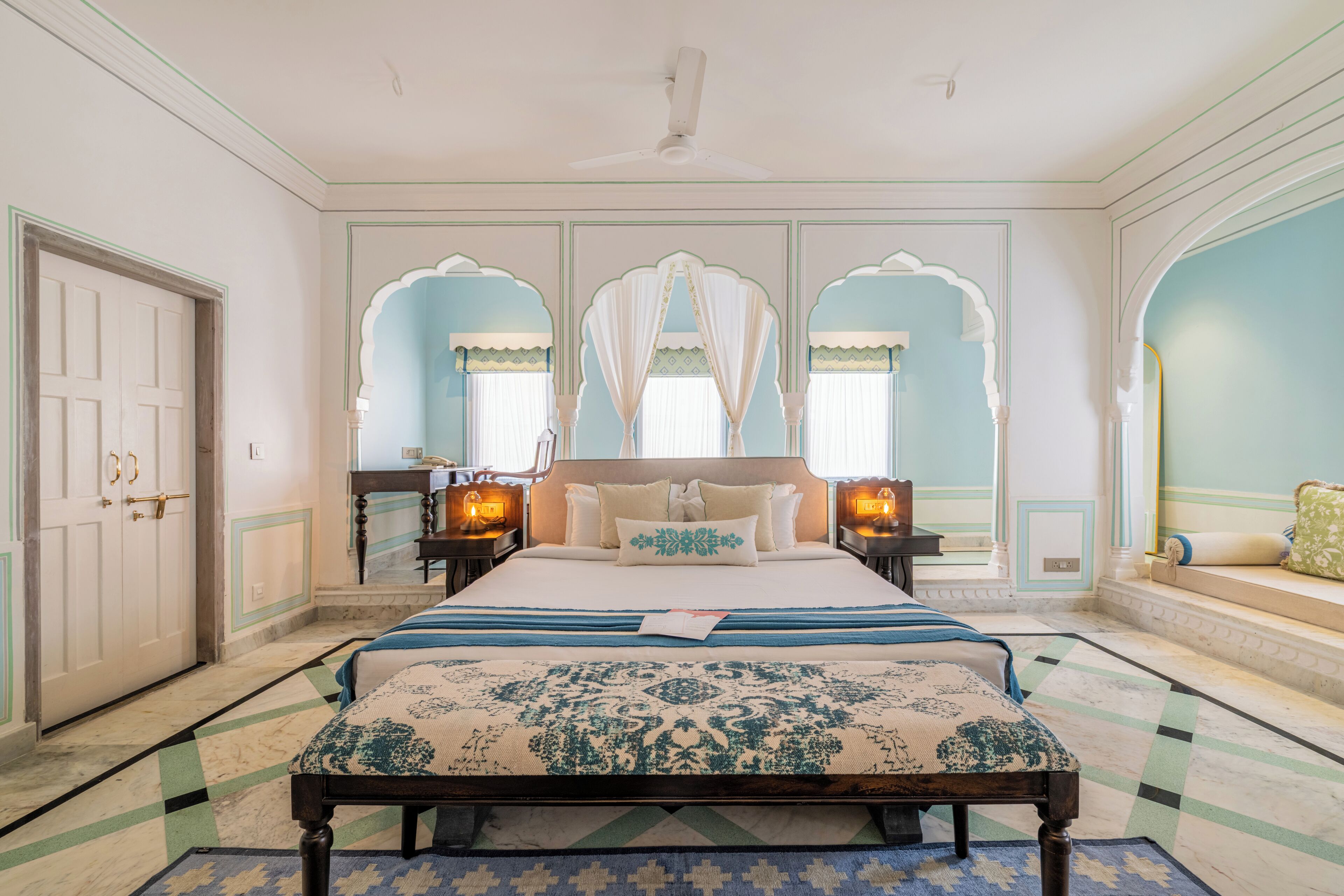 A bedroom with a large bed, two bedside tables, a ceiling fan, and a view of the outside through an arched window.