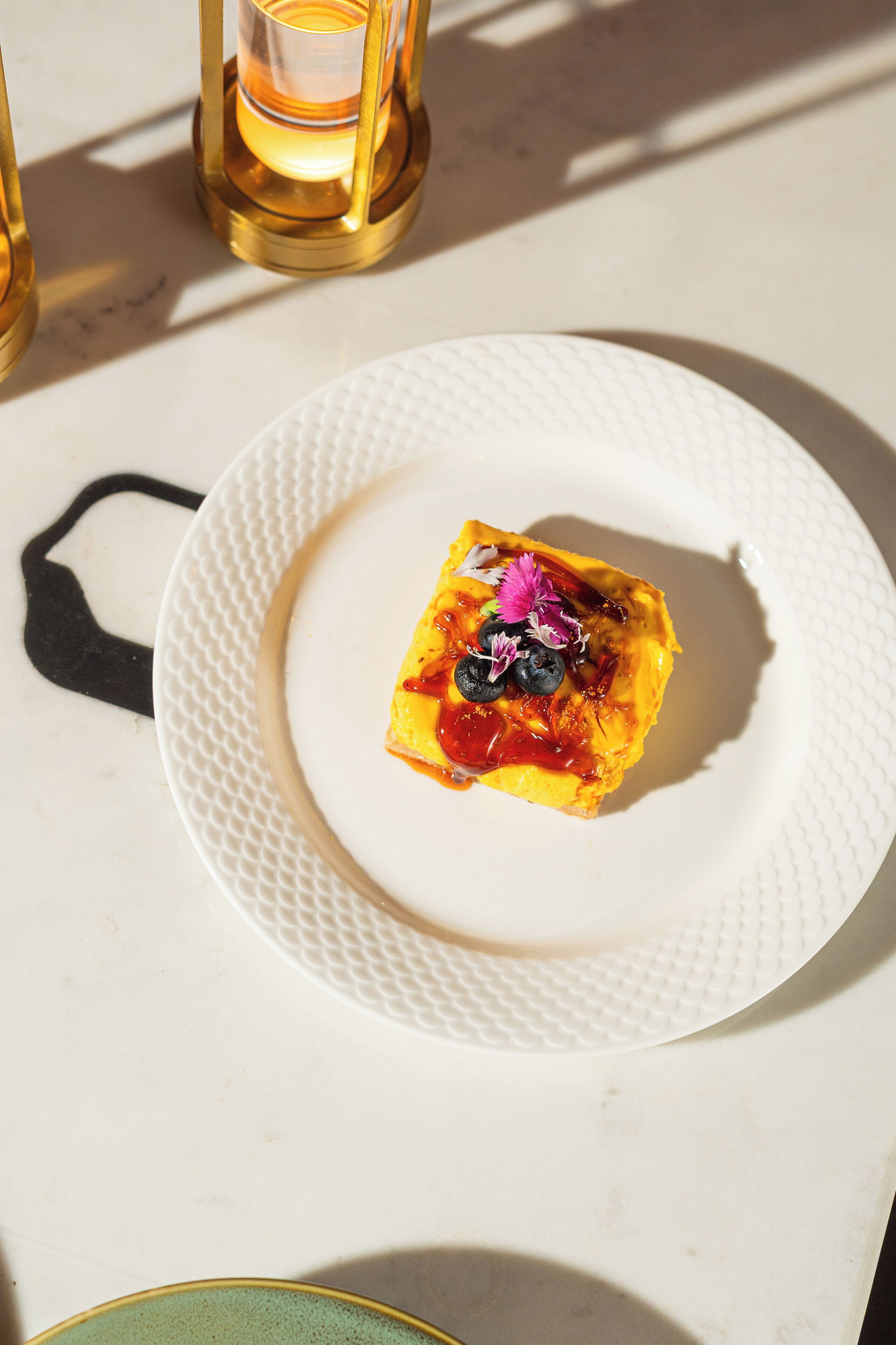 Ein weißer Teller mit einem quadratischen Nahrungsmittel, verziert mit Blaubeeren und Blumen, steht auf einem weißen Tisch, im Hintergrund eine goldene Kerze.