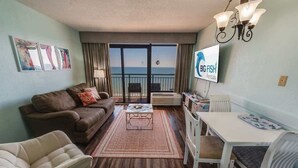 Sala de estar con sofá, sillón, mesa de centro y mesa de comedor. Hay un televisor montado en la pared y un balcón con vista al océano.